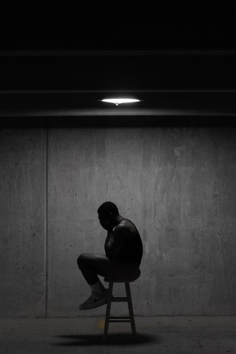 black and white photo of person with head in hands sitting on stool in dark room with single light overhead