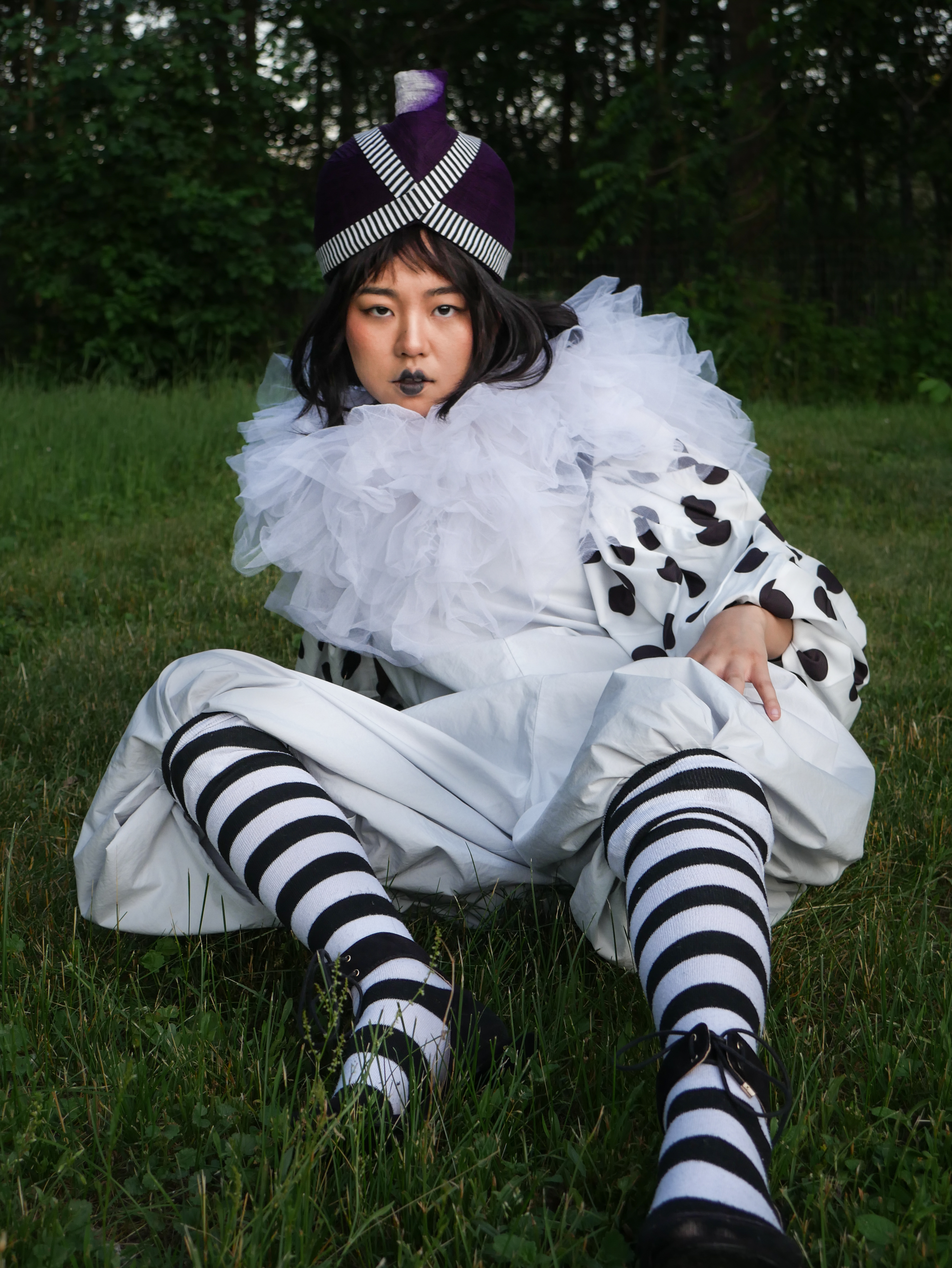colorful photo of person of Asian descent sitting on grass in black and white striped and polka dot outfit with tulle collar and headpiece