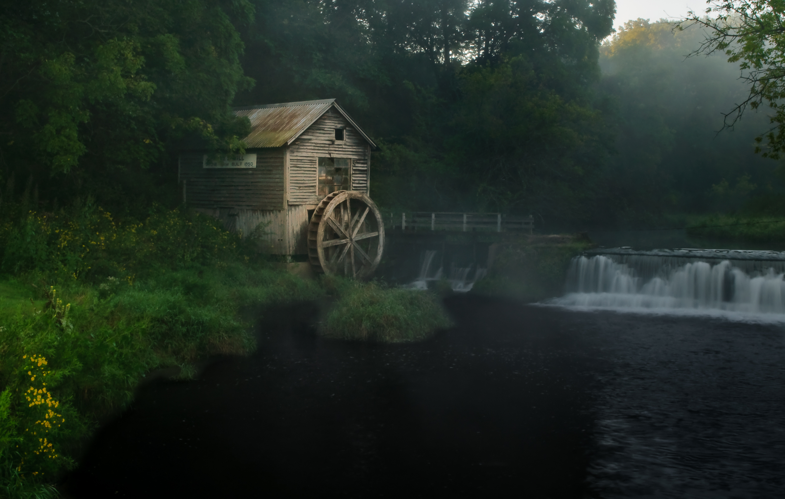 photograph of old wooden water wheel mill and dam in low light with thick green foliage and trees surrounding it