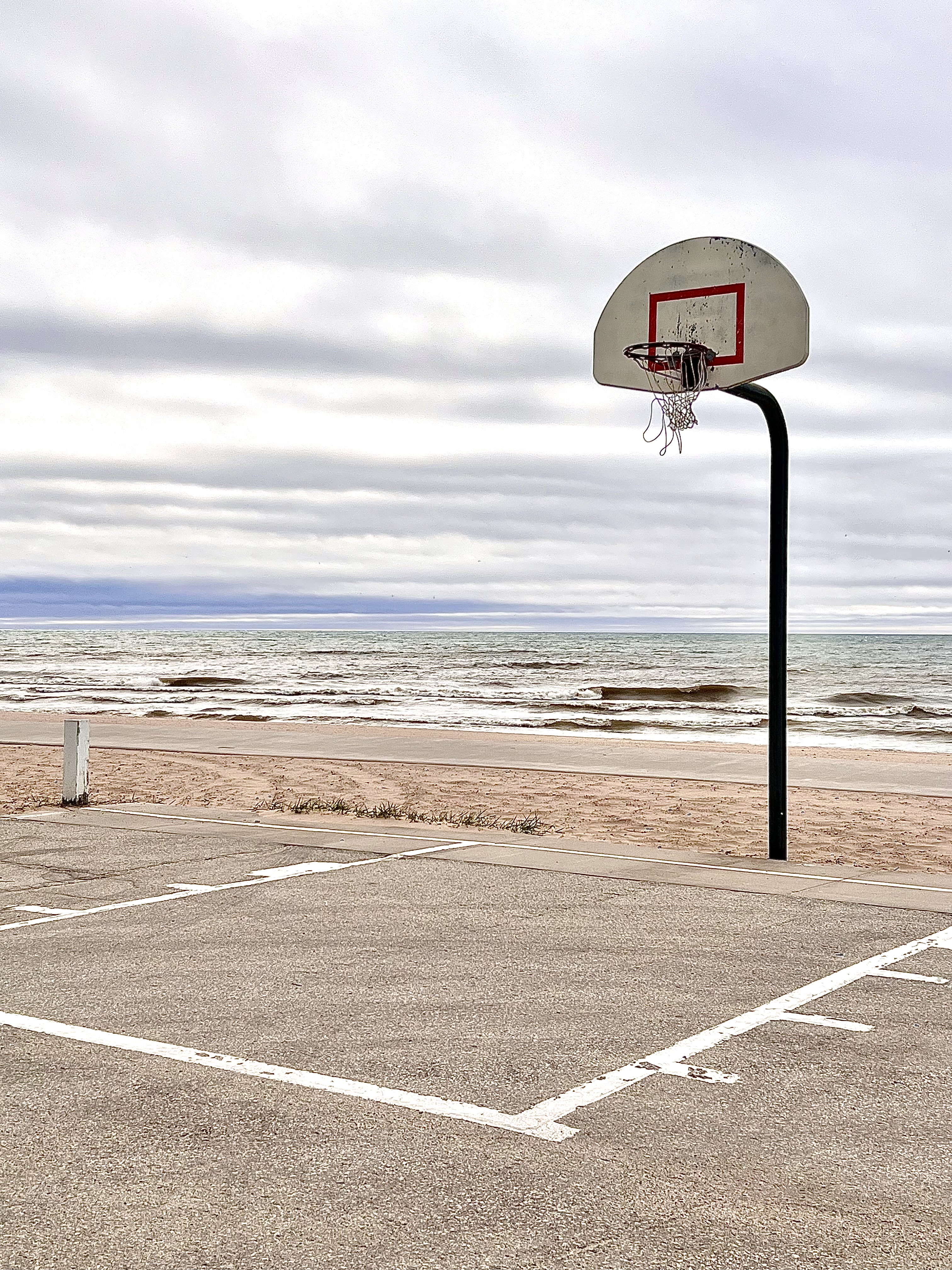 color photograph of basketball net over basketball court with water body and cloudy skyline behind it