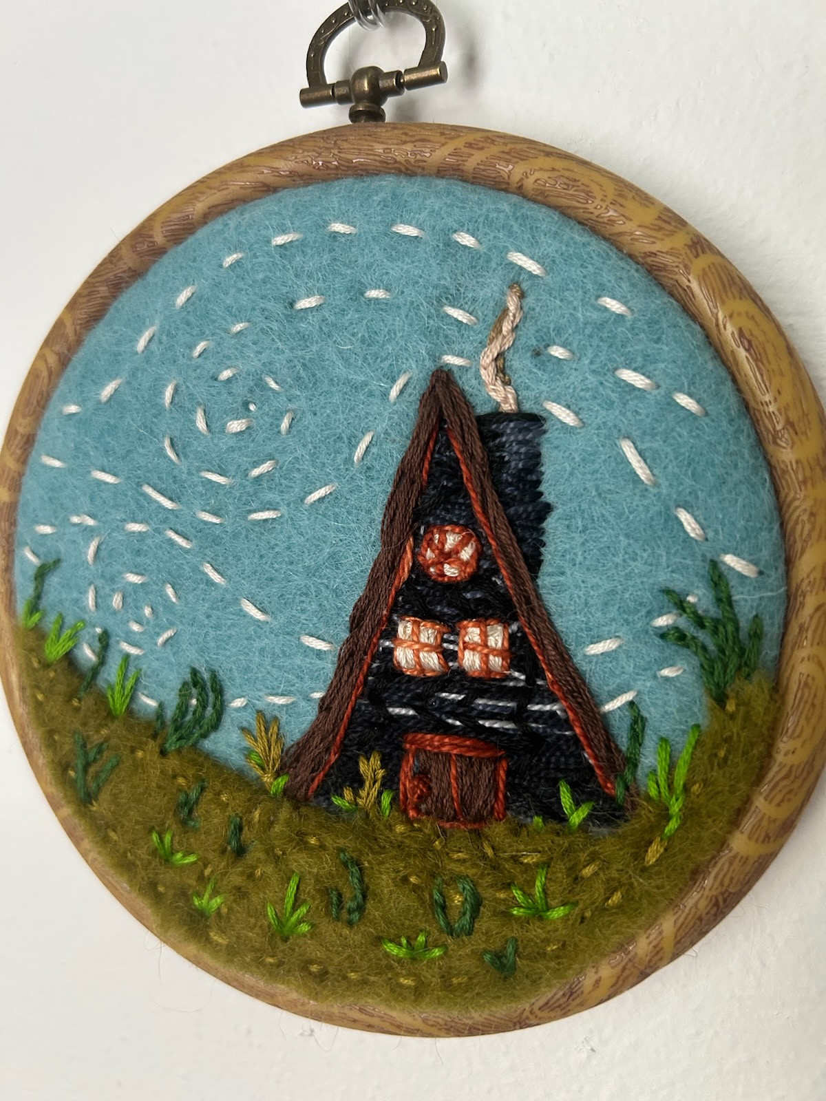 embroidered hoop of a-frame cabin with smoke coming out of chimney against a blue sky with green foliage in front