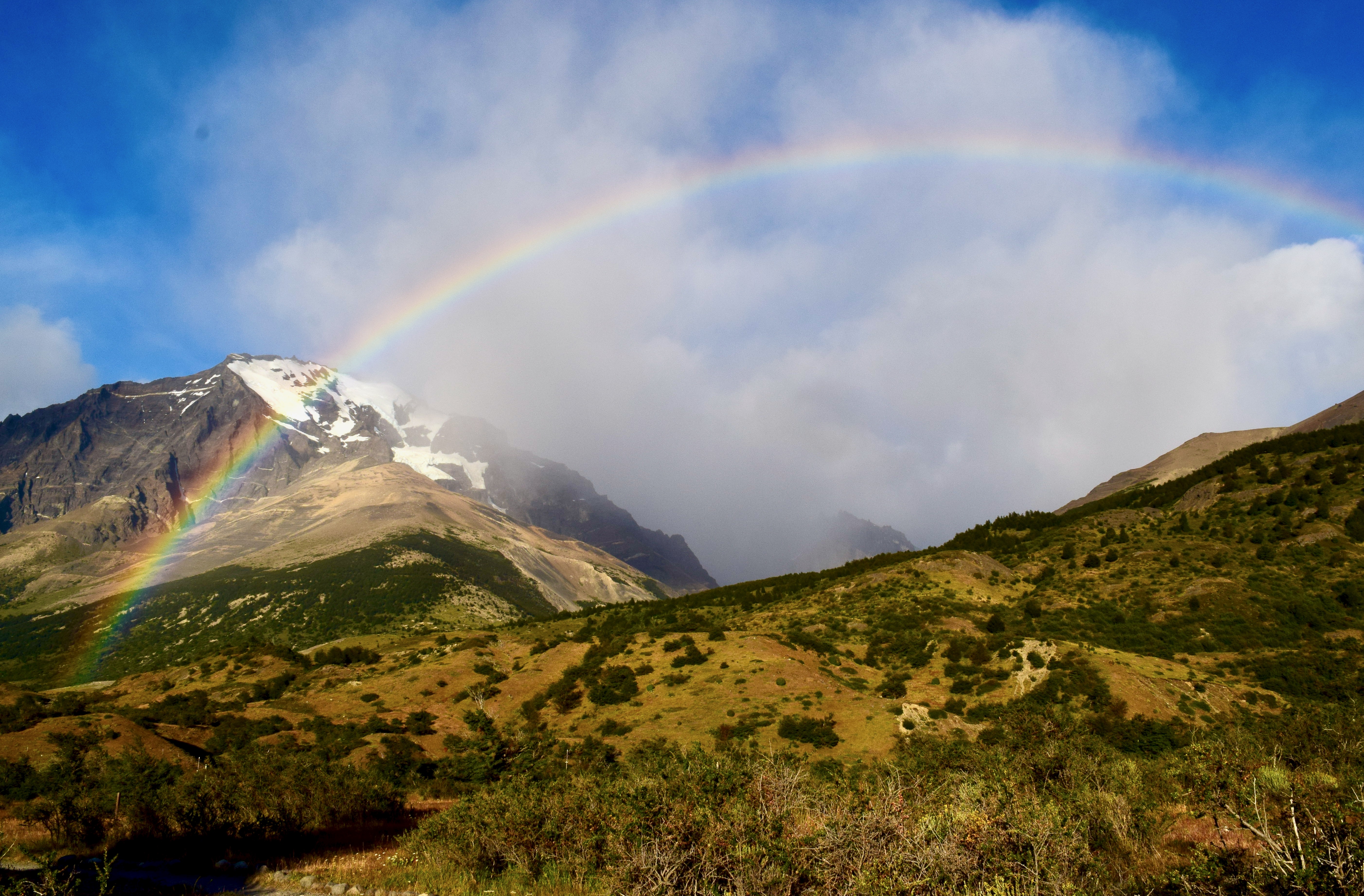 Snow-covered mountains against a blue, somewhat clear and somewhat cloudy sky with a rainbow above the valley of scrub plants
