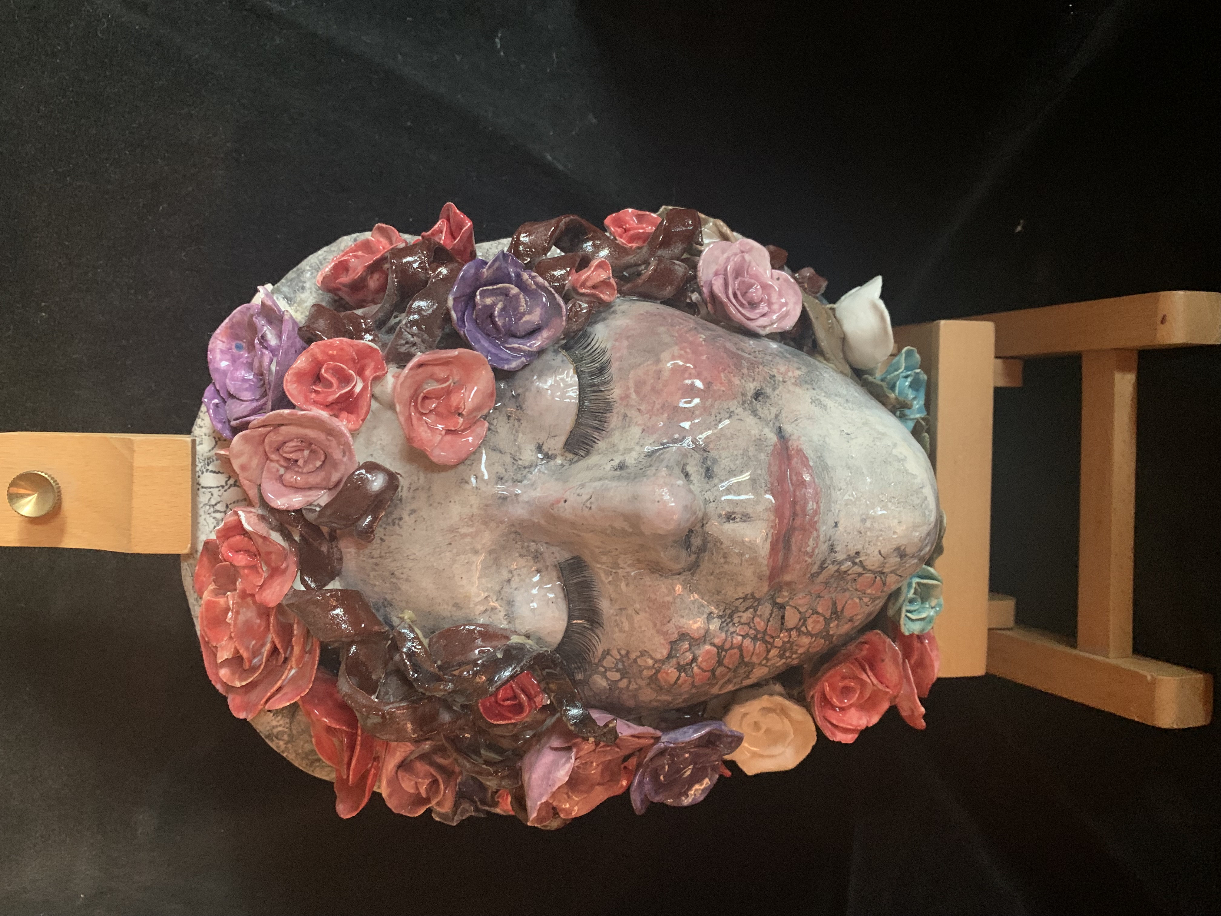 Porcelain face with closed eyes and red, purple, pink, brown flowers surrounding it sitting on a small pedestal with black background