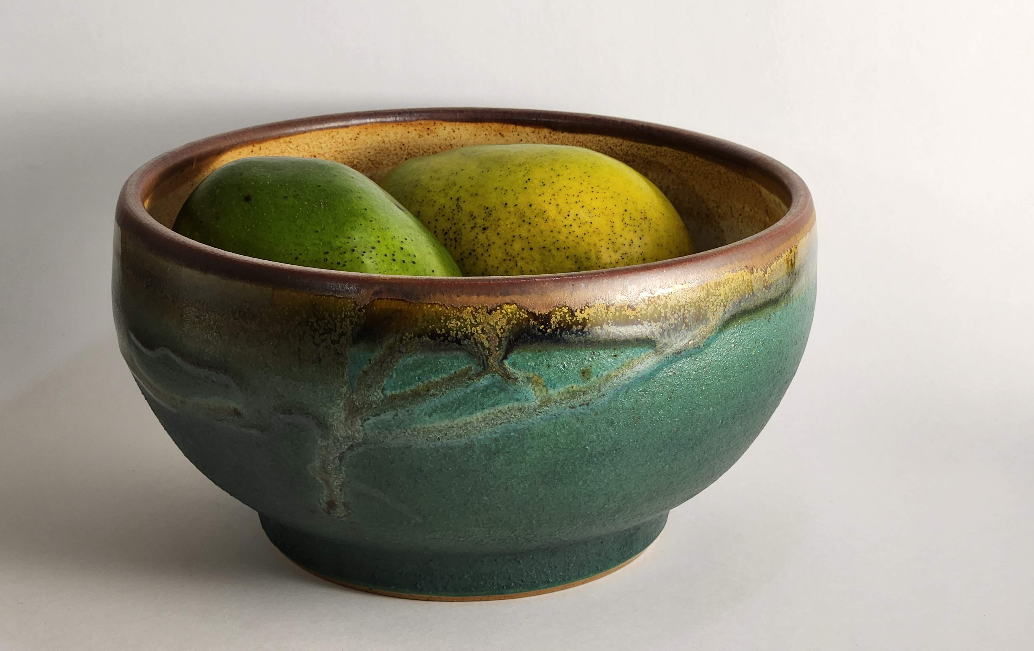 photograph of bowl with teal-green glaze outside and tan-brown glaze inside and lip of bowl, with two mangoes sitting inside bowl, photographed against a white-gray background