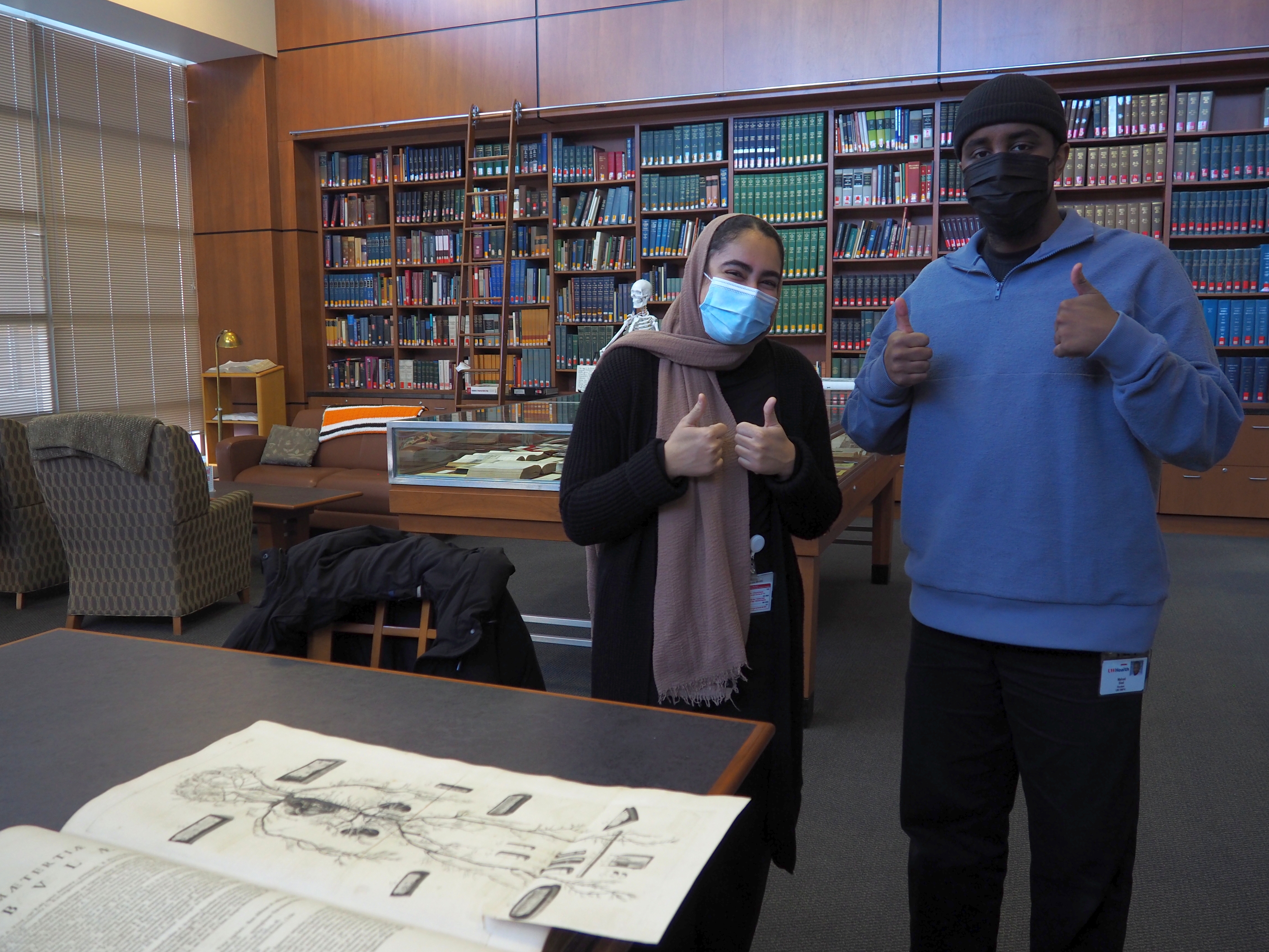 two masked medical students pose in the Ebling Library with book shelves behind and open book in front of them
